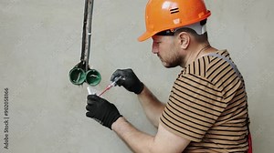 Electrician male in uniform, protective gloves and helmet checks presence of electrical voltage in socket phase uses electrical tester screwdriver.Examining wires in outlet by voltage detector indoor.