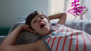 Young boy yawning with arms stretched behind his head while lying on a sofa, expressing drowsiness in the morning light, surrounded by cozy cushions and a relaxed indoor setting