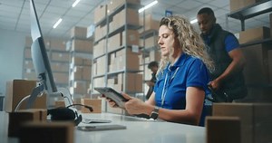 Female warehouse clerk scans parcels using tablet computer preparing packages for shipping to clients. Sorting center employees carrying cardboard boxes. Warehouse of post office or online store.