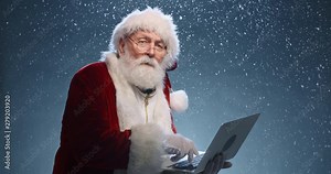 Santa Claus holding laptop in hands, typing, then looking at camera and winking, isolated over blue background - christmas spirit concept close up 4k footage
