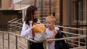 Small schoolchildren stand on a ramp with a railing near the school entrance and look into each other's open backpacks.