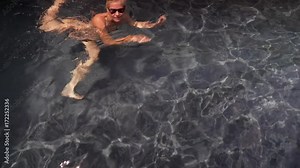 Beautiful, slim, caucasian woman wearing string bikini getting into private pool lined with flowers and grey wall. Camera moves with swimmer. Stock Video