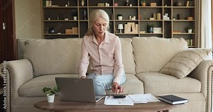 Senior female pensioner skilled online billing system user pay utility bills remotely from home office using laptop check sums on calculator. Retired hoary lady manage domestic finances analyze costs