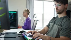 Front closeup view of young hip businessman in casual attire working on keyboard and widescreen computer in home office with woman working on another computer in the background.