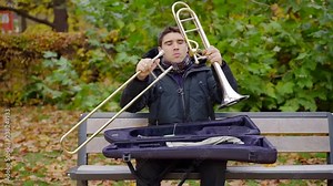 Guy assembling a trombone sitting on bench in a park.