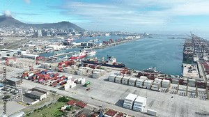 Wide aerial view of Cape Town's container terminal port, showing container stacks, both dry and reefer, in foreground. Table Mountain and Cape Town city in background.