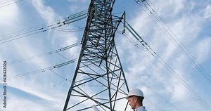 An engineer in a white protective helmet with a blueprint poster stands next to a high-voltage power line pole and looks at the poster. The engineer appears in the frame gradually.