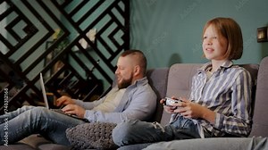 Freelancer working remotely, kid playing video game. Daughter having fun and playing a console game while dad works on a laptop