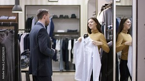 A side view panoramic filming of a clothing shop consultant offering a customer to try on a white shirt and purchase it