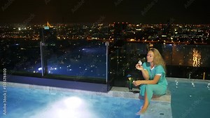 The pretty business woman spends time in a luxury hotel. The woman relaxing on the edge of the rooftop illuminated pool and drinking cocktail. Resting travel in a modern night big city.