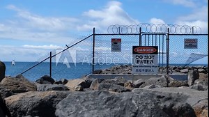 A chain-link fence with barbed wire on a breakwater in Werribee South displays multiple warning signs, including "Danger" "Do Not Enter" in multiple languages, to restrict access and warn the public