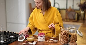 Young woman in yellow bathrobe making breakfast with a yogurt and raspberry, decorated meal in the glass bowl. Healthy vegan cereal meal for breakfast concept