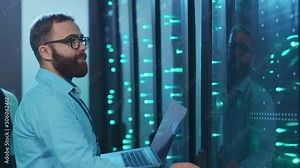 Female IT server specialist cooperating in digital high tech room of data center. Bearded administrator opening rack server updating hardware networking with laptop computer. Teamwork.