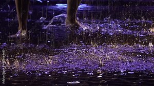 Contemporary dance ballerina dances sensually in rain on water surface, creating splashes. Silhouette of wet woman in white shirt move plastically in dark studio with blue light. Slow motion. Close up