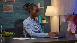 Black woman typing on computer and writing list of tasks on sticky notes putting it on laptop sitting on desk in living room workplace late at night. African freelancer working from home overtime