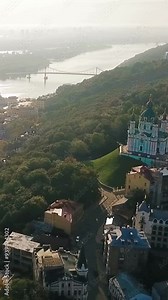 aerial vertical video of Kyiv capital of Ukraine. camera flies over st. andrew church, green Zamkova Hora hill and approaches the Dnipro river and pedestrian bridge. Morning sunrise soft sun backlight