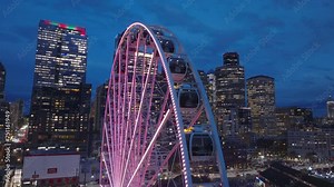 Colorful illumination of Seattle Great Wheel in amusement park on pier in Waterfront Park. Cinematic aerial with panorama of Seattle at night. Beautiful skyscrapers in modern city downtown.USA tourism