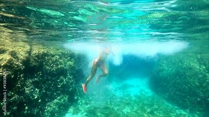Underwater video. Young girl jumping off a cliff into a blue clear sea. Crystal clear water splashing as she dives off a rough rocky shore underwater, you can see her swimming underwater