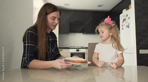 Mum and daughter cooking in the kitchen. Woman showing her daughter nice cake on a plate, girl in pink bow being happy and pointing at it/touching it