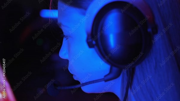 A close-up of a streamer girl playing an online game on a computer in a computer club, she talks to the participants and her face is illuminated by the light from the screen. Side view. Live streaming