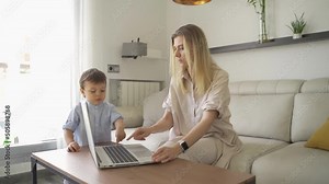 blonde woman typing on laptop shows boy how to use computer. playing and education of the basics. future programmer. bright living room. Slow motion video footage. Working mother at home with toddler