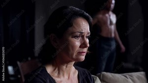 A woman with a frightened face sitting on a sofa. Behind her a man altered and violent