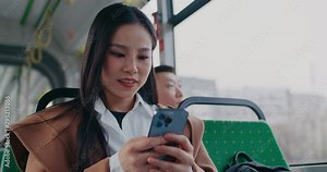 Beautiful Chinese woman in white shirt and yellow jacket traveling to work or university. Typing message on smartphone. Smiling after positive response. Other passengers in blurred background.