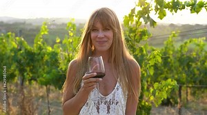 Adult blond woman drinking red wine during sunset time with wineyard in background