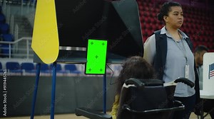 Female voter, woman with SMA disability in wheelchair votes in voting booth at polling station. Tablet computer for voting with displayed chroma key green screen. United States Presidential Elections.