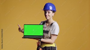 Female construction worker holding laptop with greenscreen in studio, pointing at chroma key display with blank copyspace. Using isolated mockup template on computer, repair work.