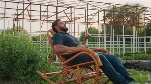 Gardener seated on wooden rocker chair holding clear mug of herbal tea leaning back gazing into distant garden under warm sunset light surrounded by leafy plants metal trellis | Premium Stock Video Footage