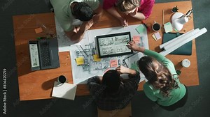 Top down view of civil engineers using tablet to design house structure while sitting at table with project plan placed. Aerial view of team planning and sharing idea about construction. Symposium.