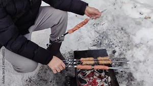 A man cooks outdoors in winter, grilling kebabs on skewers over coals in a barbecue.
