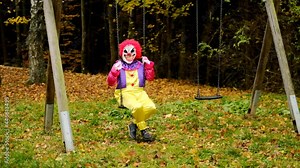 Halloween. Evil clown costume. Sinister smiling clown swinging on a swing on a playground in an autumn park.Horror and fear