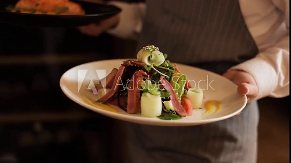 Professional female waiter dressed in elegant apron carrying plates with fancy dishes in both hands for guests of restaurant. Excellent service and beautiful presentation of cooking masterpieces.