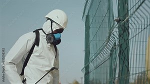 African American ecologist in protective coverall, hardhat and respiratory mask using backpack sprayer while applying chemicals to fence by gas pipeline outdoors