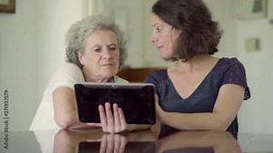 Two women sitting at table and using tablet. Adult daughter showing how to use tablet her senior mother. Technology concept