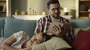 Slow-motion shot of a handsome young man sitting on the sofa, telling story from the book to his wife who is lying on his lap