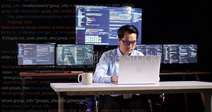 Asian Male Programmer Writing Code By A Laptop Using Multiple Monitors Showing Database On Terminal Window Desktops In The Office. Code Animation Footage
