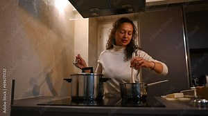 Beautiful Latin American woman standing by kitchen stove and mixing ingredients in saucepan, enjoying preparing Italian pasta for dinner at cozy home interior with romantic lighting in the evening.