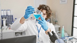 Curly-haired man in laboratory examines blue chemical in flask with microscope and equipment around.
