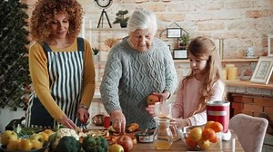 Multi-generational family is having fun making fresh fruit juice in the kitchen. Grandma, daughter and granddaughter put pieces of fruit in an electric blender, preparing healthy fruit smoothie