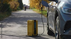 Using of home-invented portable battery to charge electric car out of the city. Light blinks indicating its work. Peaceful sunny autumn day, bright yellow leaves and road background. High quality 4k