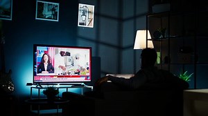 At night, young guy follows latest headlines on tv relaxing on couch in the living room, watching news program. On international network person pays attention to information broadcasted. Camera A.