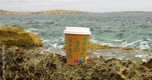Plastic cup with cover with Save Our Seas phrase on rock at sea waves closeup. Warning about sea protection importance. Environment conservation