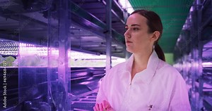 Portrait of a Female Agricultural Engineer Walking in a Vertical Farm Facility Next to Racks with Fresh Natural Crops. Hydroponics Specialist Using a Tablet Computer, Studying and Cultivating Plants