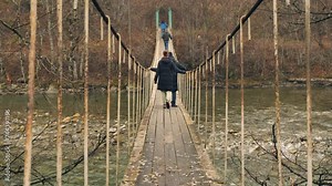 Crossing a suspended wooden bridge over a river. A group of people walking across a bridge over a mountain river. View from behind. Hiking. Active lifestyle
