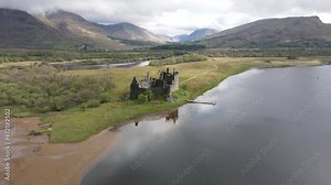 Kilchurn Castle, an old Scottish fortress mirrored on the peaceful waters of Loch Awe. Aerial drone reveals the historic stone ruins against a scenic cloudscape. Panoramic shot