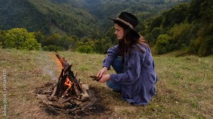 A beautiful young woman in hat, kindles campfire in mountains. Traveler girl, sitting near to campfire. Explore travel real wilderness lifestyle