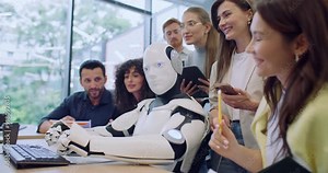 Side view of humanoid robot typing on keyboard while sitting at desk in front of big monitor. Students around noting everything and discussing their observation. Studying engineering or programming.